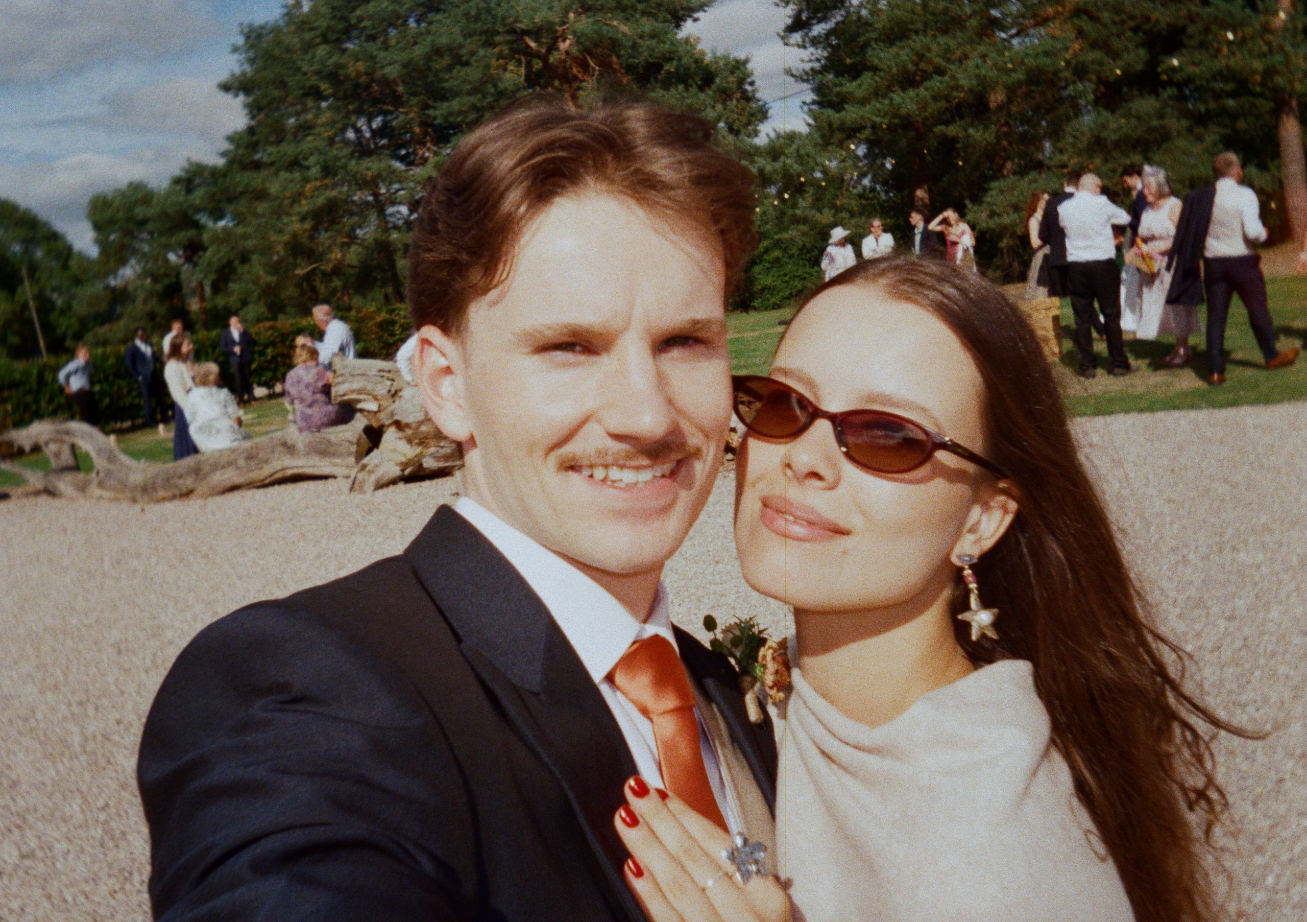Film photo of a wedding couple taking a selfie, him in a suit, her in oval sunglasses and star earrings