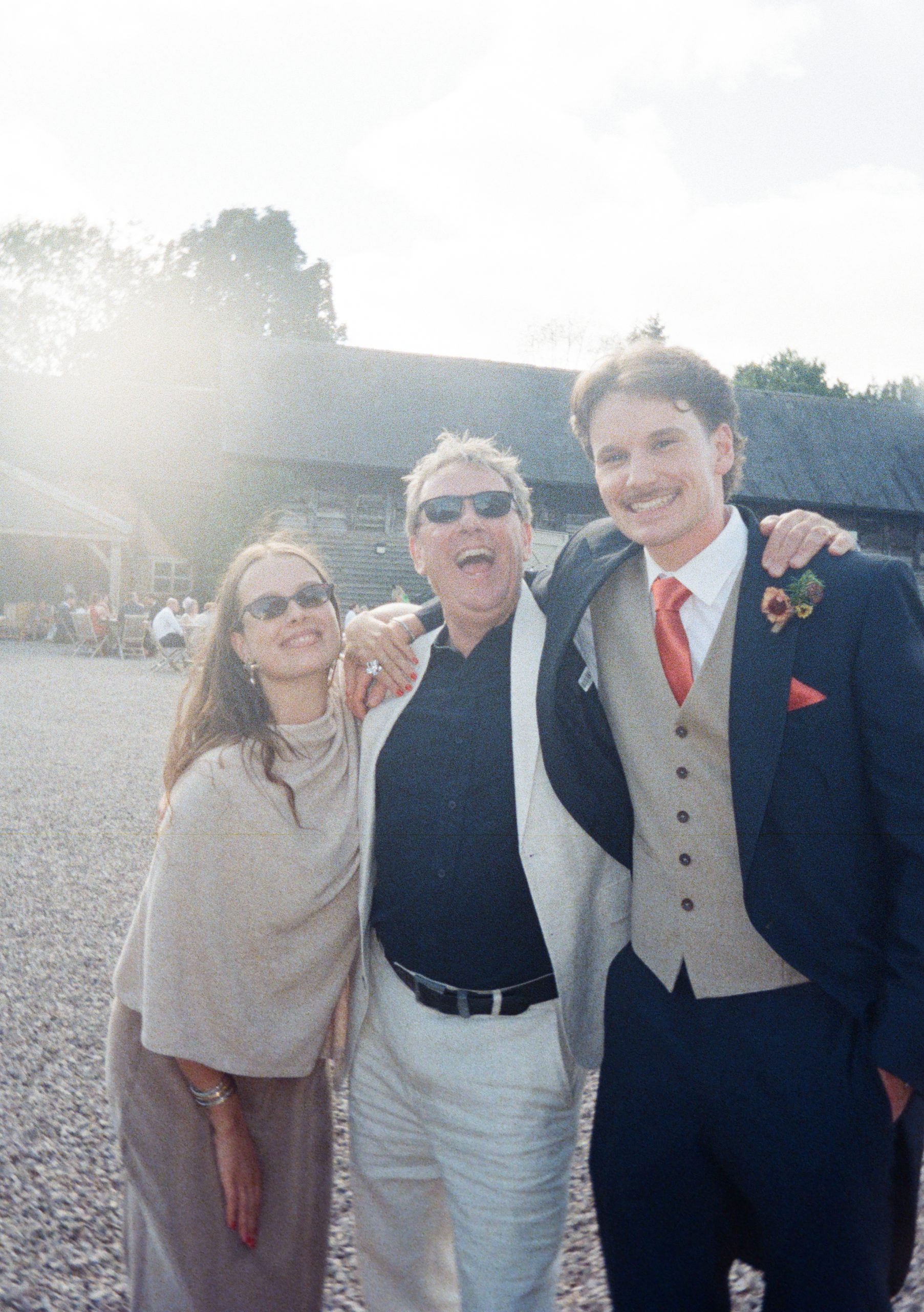 Film photo of three wedding guests arm in arm outside, one in sunglasses and a white jacket