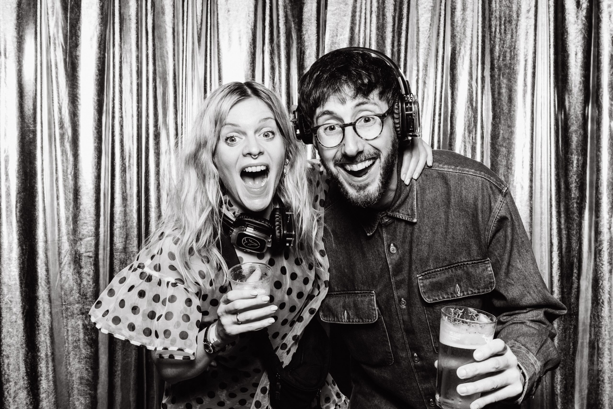Black and white booth print of a woman in a polka dot top and a man in glasses and headphones, both cheering at the camera